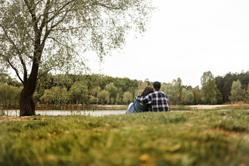 Young couple in love sitting on the grass near the lake and hugging. Rear View of Young Couple Sitting on grass in the beautiful spring day.