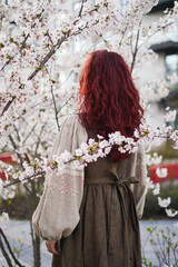 Women stand back with long red hair in traditional Ukrainian clothes - beige linen shirt embroidered with a cross. blooming trees and a woman in an embroidered shirt