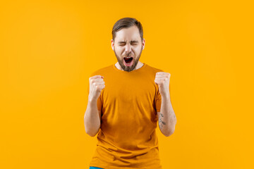 Studio shot of emotional soccer supporter man cheering for his favourite team making winners gesture clenching his fists, isolated over bright colored orange yellow background