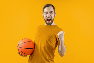 Studio portrait of young attractive basketball player or supporter posing over bright colored orange yellow background holding the ball in hand and making winner's gesture clenching his fist - Powered by Adobe
