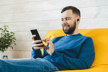 Portrait of young handsome casually dressed entrepreneur man using mobile phone for a video call with his client while sitting on comfortable yellow sofa at home