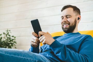 Close up portrait of handsome smiling young man listening music via wireless earphones and texting message on his smartphone