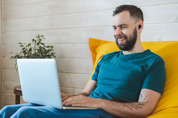 Indoor shot of handsome smiling bearded young man having video-call on his laptop while sitting on comfortable yellow sofa at home