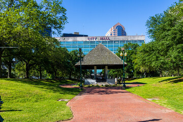 a beautiful spring landscape at Duncan Plaza with a red brick footpath, lush green trees, grass and plants, a pergola, and the City Hall Building and skyscrapers in the skyline in New Orleans
