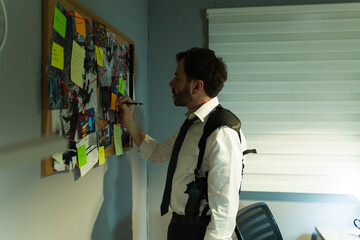 Serious Caucasian detective examines evidence and notes pinned on a board, connecting the dots on a...
