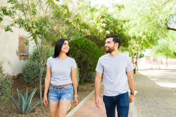 Happy young hispanic couple strolls in casual clothing, showcasing blank t-shirt mockups, in a sunny outdoor environment