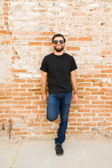 Smiling hispanic man in black t-shirt and jeans posing against a rustic brick wall for a mock-up...