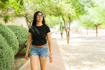 Confident Hispanic woman walking in a sunny park wearing a plain black t-shirt for mockup designs