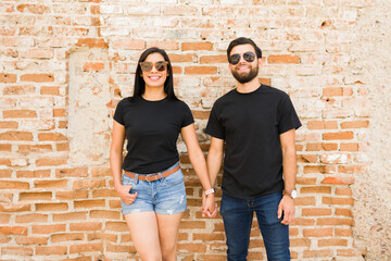 Happy hispanic couple in black t-shirts holding hands, perfect for brand mockups, standing in front of a weathered brick wall
