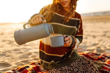 Happy woman in a stylish sweater sits at picnic on the beach drinks a hot drink from a thermos and enjoys nature. Travel, weekend, relax and lifestyle concept.