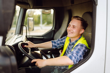 Obraz premium Handsome driver at the wheel of a truck at work. Driver or forwarder on truck and trailer, on a transshipment point. Transportation service.