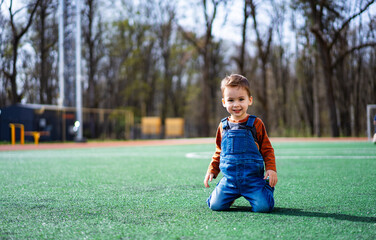 A young child is sitting on the grass in a park. The child is wearing a blue overalls and a brown shirt.