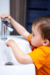 A young child is playing with a faucet in a sink. The child is wearing an orange shirt