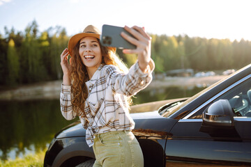 Young female tourist traveling  by car taking selfie standing next to the car. Active lifestyle,...