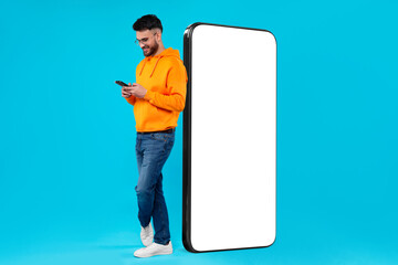 Man with mobile phone standing near huge device with empty screen on light blue background. Mockup...
