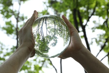 Overturned reflection of beautiful green trees outdoors, low angle view. Man holding crystal ball...