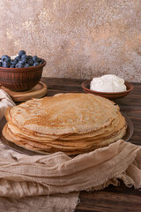 A plate of golden pancakes topped with blueberries, accompanied by a bowl of sour cream, a portion of fresh cottage cheese and a glass of milk. On a beige-golden background with a green runner.