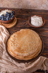 A plate of golden pancakes topped with blueberries, accompanied by a bowl of sour cream, a portion of fresh cottage cheese and a glass of milk. On a beige-golden background with a green runner.