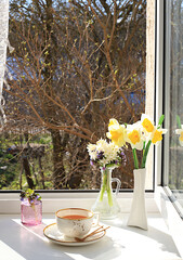 Cup of tea,croissant and spring flowers in a vase on a sunny window.The concept of home comfort and greeting of spring.Beautiful flower arrangement with hyacinths and daffodils,selective focus.