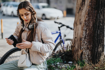 A young woman checks her phone with her headphones around her neck, sitting beside her bicycle in the city.
