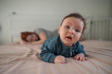 A three-month-old boy lies on his stomach on the bed and his mother sleeps behind him. Postpartum depression.