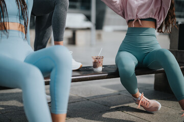 Close-up of two fit women in active wear resting on a bench with healthy smoothies outdoors.