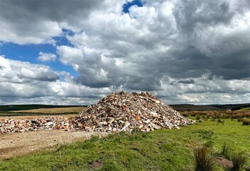A large pile of rubble is prominently situated in the center of a grassy field under a dramatic sky filled with clouds near, Sutton-in-Craven, UK