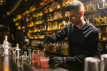 Skilled barman makes refreshing cocktails for pub guests. Bartender put ice to cool beverage using tongs standing background shelves with different bottles of alcohol