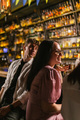 Young people spend time together in trendy club. Brunette guy and girl watch other visitors to bar...