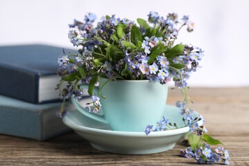 Beautiful forget-me-not flowers in cup, saucer and books on wooden table against light background, closeup