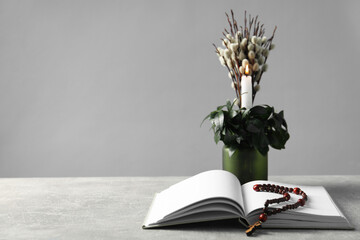 Burning candle, plant with willow branches, Bible and rosary beads on grey table, space for text