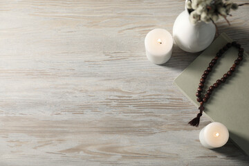 Rosary beads, burning candles and book on wooden table, flat lay. Space for text