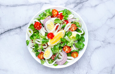 Fresh salad with squid, eggs, cherry tomatoes, onion, lamb lettuce and olive oil with lemon juice dressing, marble table background, top view