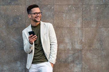Smiling businessman with smart phone. Handsome executive looking away while standing against wall....