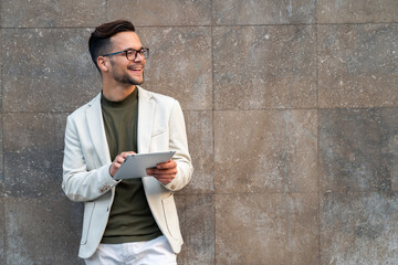Young happy businessman wearing suit and eyeglasses using digital tablet against a building wall.