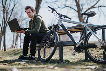 A good looking young man uses his laptop while sitting on a bench in a park, with his bicycle...