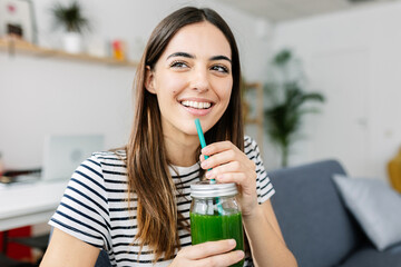 Cheerful young millennial woman enjoying homemade green smoothie juice at home. Diet and healthy lifestyle concept.