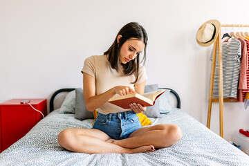 Young beautiful woman reading book while sitting on bed at home. Leisure activity concept.