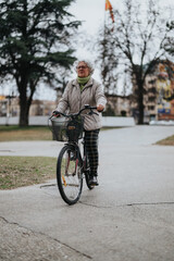 An elderly woman in casual wear rides her bike on a path with trees and a flag in the background.