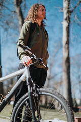 A young man stands casually with his bicycle in a park, looking thoughtfully away, enjoying the warm sunshine and fresh air.