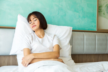 A Thai woman wearing a white T-shirt is asleep while sitting on a white bed in a room with light shining through the window.