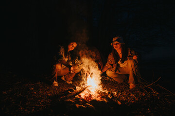 Two friends experiencing the warmth and joy of a campfire outdoors at night, preparing food and...