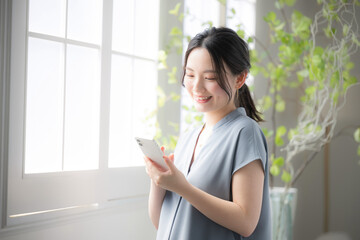 窓辺でスマホを持つ笑顔の若い女性　スマホで検索や探す画面を見る