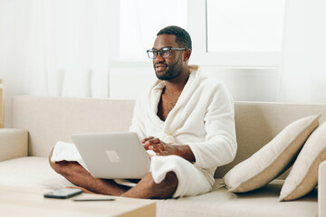 Smiling African American Man in Bathrobe Typing on Laptop while Working from Home on a Modern Sofa...