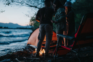 A group of friends gathers around a campfire by a lakeside at night, surrounded by nature, sharing...