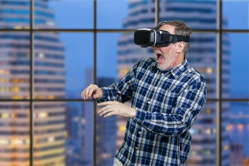 Portrait of an elder man in vr headset with his mouth open. Checkered window background with night cityscape view.