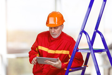 Happy male factory manager using digital tablet in warehouse. Worker wearing red robe and orange helmet.