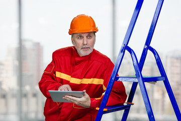 Male elder contruction worker with tablet pc. Aged man with red helmet leans on a blue ladder. Indoor window background.
