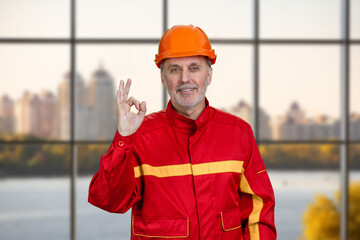 Happy smiling construction worker in red uniform shows okay gesture sign. Checkered window background.