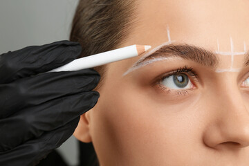Beautician preparing young woman for procedure of permanent eyebrow makeup on grey background,...